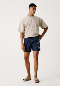 T-shirt oversize beige, pantaloncini da bagno blu navy con testo bianco; sandali casual; tessuto morbido, vestibilità comoda e cintura elastica.