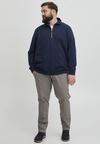 Námornícky pulóver na zips vyrobený z mäkkého materiálu s vysokým golierom, v kombinácii so svetlo šedými nohavicami a čiernymi topánkami. Jednoduchý, ležérny dizajn.