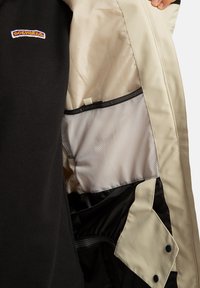 Beżowa kurtka rozpięta, aby pokazać wnętrze z białą siatkową kieszenią i czarną podszewką, trzymana w ręku; osoba nosi czarną koszulę z logo DGSHOECO.