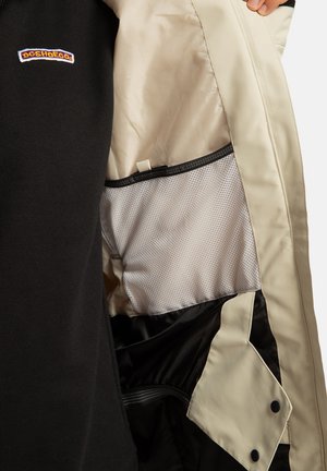 Beige jas opengeklapt om de binnenkant te tonen met een witte gaaszak en zwarte voering, vastgehouden in de hand; persoon draagt een zwart shirt met het DGSHOECO-logo.