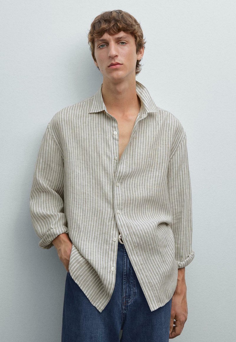 Helles beige Leinenhemd mit vertikalen weißen Streifen, Kragen und Knopfleiste. Figurbetonte, an den Bündchen hochgerollte Ärmel. Dunkelblaue Jeans.