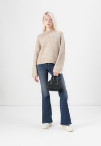 Beige gestrickter Pullover mit langen Ärmeln, kombiniert mit blauen Schlagjeans. Das Model hält eine kleine schwarze strukturierte Handtasche und trägt weiße Sneaker.