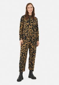 Løvepræget jumpsuit i brun og sort, lange ærmer, afslappet pasform, lavet af blødt stof, parret med sorte ankelstøvler.