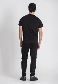 Camiseta de algodón negra de manga corta y cuello redondo, combinada con jeans negros y zapatillas negras. Vista desde atrás.