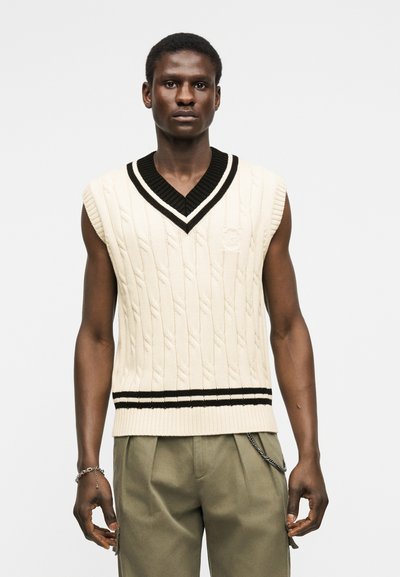 Άνδρας που φορά αμάνικο κρεμ γιλέκο με πλεκτό σχέδιο κοτσίδα, με μαύρο περιθώριο σε σχήμα V στο λαιμό και πράσινο-μπεζ παντελόνι, μπροστά σε λευκό φόντο.