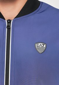 Lengvas bombonešis džemperis karališkai mėlynos spalvos, su juodu užtrauktuku ir raštuotu apykakle. Pridedamas juodas ir baltas logotipo ženklelis ant krūtinės.