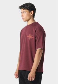 Nuori mies, jolla on yllään viininpunainen lyhythihainen paita, jossa on pieni oranssi graafinen kuvio rinnassa, seisoo profiilissa vaaleaa taustaa vasten.