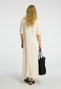 Vestido maxi color crema con mangas cortas, que presenta detalles de costura diagonal. La modelo lleva un bolso de mano negro tejido y sandalias negras.