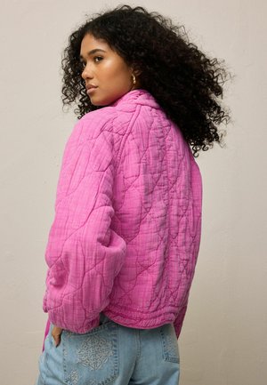 Frau mit lockigem Haar, die eine leuchtend pinke gesteppte Jacke und hellblaue bestickte Jeans trägt und über die Schulter blickt.