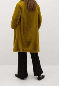 Manteau en polaire jaune moutarde, coupe oversize avec col. Porté avec un pantalon large texturé noir et des ballerines noires.