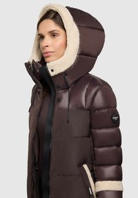 Giacca a vento marrone con cappuccio foderato in pelliccia, dotata di zip frontale, dettagli con cordino e patch con logo sulla manica.