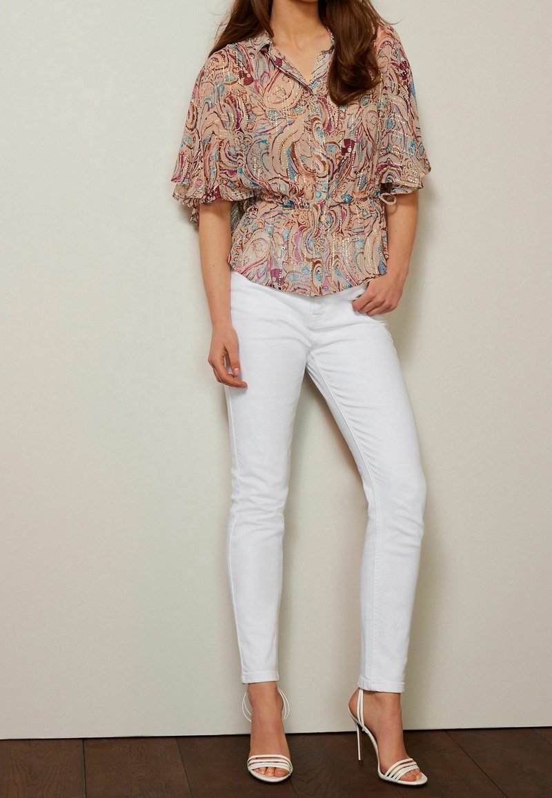 Femme portant un chemisier à manches courtes à motifs, un pantalon blanc ajusté et des sandales blanches à talons hauts, debout contre un mur uni.