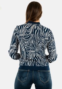 Giacca bomber blu navy con motivo di foglie tropicali bianche, polsini e orlo a costine, caratterizzata da un dettaglio con zip sul retro.