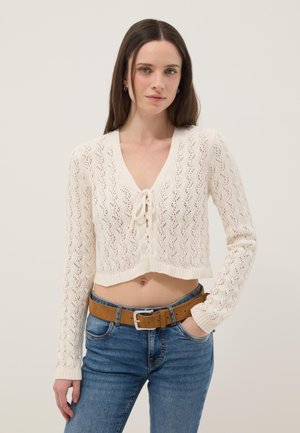 Giovane donna con lunghi capelli castani che indossa un cardigan corto lavorato a maglia color crema con fiocco frontale e jeans blu con cintura marrone.