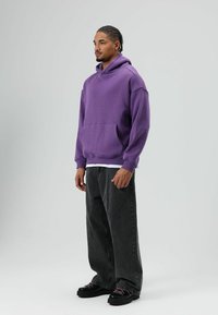 Sweat à capuche violet avec une poche frontale, coupe ample, poignets côtelés, porté sur une chemise blanche ; assorti à un jean large gris foncé et des chaussures noires.