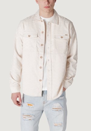 Camicia a maniche lunghe con motivo crema, dotata di due tasche sul petto, texture sottile e orlo arrotondato. Indossata sopra una t-shirt bianca, abbinata a jeans strappati.