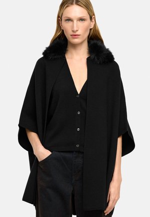 Kobieta ubrana w czarny sweter na guziki i pasującą do niego pelerynę z czarnym futrzanym kołnierzem, stojąca z jedną ręką w kieszeni.