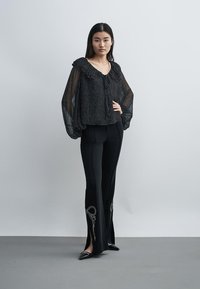 Femme debout portant un chemisier noir transparent avec des volants et un pantalon noir avec des fentes brodées, posant devant un fond uni.