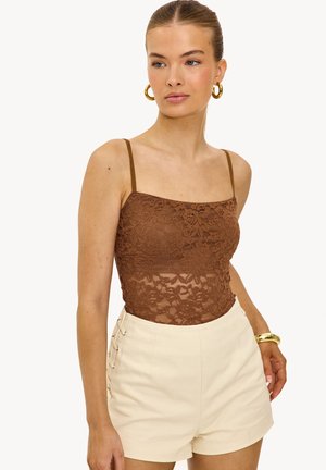 Jonge vrouw die een bruine kanten camisettop draagt, beige short met hoge taille en zijveters, gouden creolen en een gouden armband, tegen een witte achtergrond.