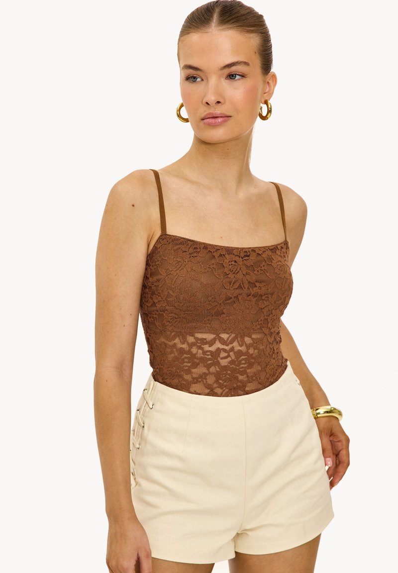 Jeune femme portant un caraco en dentelle marron, un short beige taille haute avec laçage sur le côté, des boucles d'oreilles créoles en or et un bracelet en or, sur fond blanc.