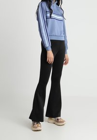 Felpa zip blu chiaro con tre strisce blu scuro sul petto, abbinata a pantaloni neri a zampa d'elefante e sneakers beige.