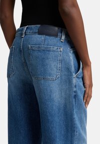 Blå denimjeans med en avslappnad passform, med bakfickor och en slät läderetikett på midjan. Texturen verkar något urblekt.