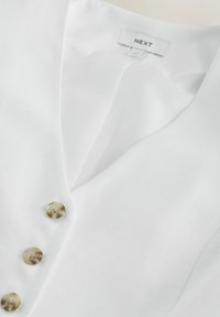 Blouse blanche à col avec des boutons beige marbrés et une étiquette de marque « NEXT » près de l'encolure.