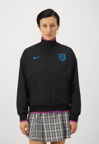 Black jacket with high collar, zip closure, front pockets, blue Nike logo, and England crest; features purple accents along cuffs and hem.