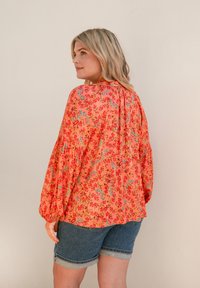 Blusa floreale in corallo con motivi multicolori, dotata di maniche lunghe a palloncino e dettagli arricciati sul retro, abbinata a pantaloni corti in denim.