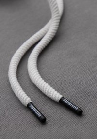 Gros plan sur les extrémités d'un lacet blanc tressé avec des embouts noirs portant le label "DIMARE" sur un fond de tissu gris texturé.