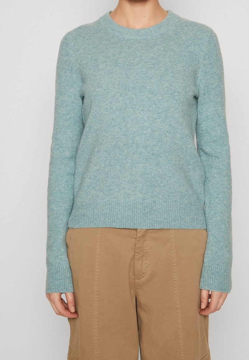 Personne portant un pull tricoté bleu clair avec des poignets et un ourlet côtelés, associé à un pantalon beige sur un fond uni pâle.