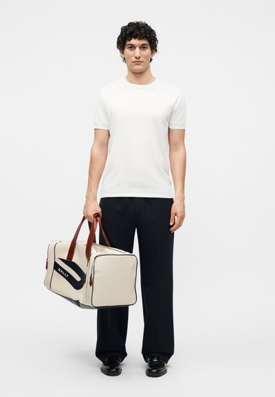 Νεαρός άνδρας με σγουρά μαλλιά, που φοράει λευκό πουκάμισο και μαύρο παντελόνι, κρατώντας μια μπεζ τσάντα ταξιδιού Bally με καφέ χερούλια.