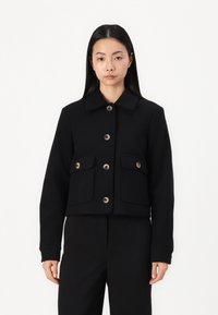 Musthave musta lühike jakk, mis on valmistatud tekstureeritud kangast. Omab krae, nööpe ja kahte esist taskut flappidega. Puhtne, minimalistlik disain.