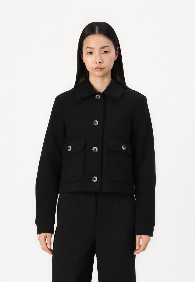 Musthave musta lühike jakk, mis on valmistatud tekstureeritud kangast. Omab krae, nööpe ja kahte esist taskut flappidega. Puhtne, minimalistlik disain.