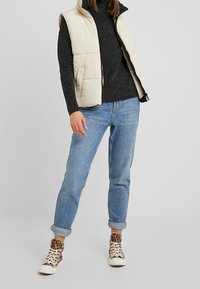 Chaleco acolchado beige sobre un suéter gris oscuro, combinado con unos vaqueros claros con puños y zapatillas beige con un diseño de estampado de leopardo.