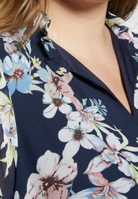 Tmavě květinová blůza s výstřihem do V a skládacím límcem; má velké světle modré a růžové květy na námořnickém pozadí. Hladká textilie.