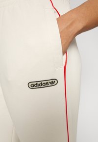 Adidas sweatpants gjorda av tyg i en krämfärg, med röda sidopaneler och en svart logotyp på framlommen.