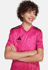 Roze sporttrui met een glanzende afwerking; heeft een geometrisch patroon, een v-halsontwerp en drie zwarte strepen op elke schouder.