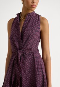 Ermeløs kjole i mørk lilla stoff med et lite geometrisk mønster i rødt og blått. Har krage og en knyttedetalj foran.