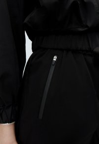 Close-up van een zwart kledingstuk met een elastische tailleband en een verticale ritszak aan de zijkant.