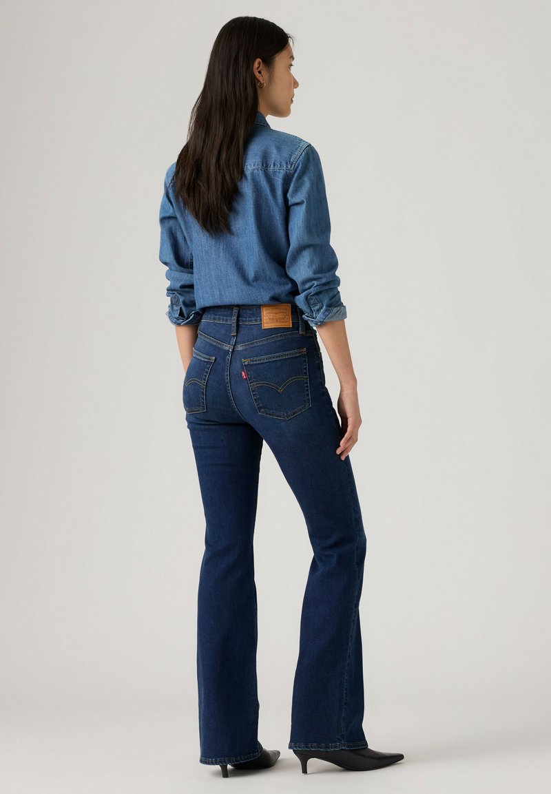 Femme debout de profil portant une chemise en jean aux manches retroussées, un jean évasé bleu foncé et des talons noirs pointus sur un fond uni.