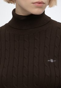 Gros plan d'une personne portant un pull à col roulé en maille torsadée marron foncé avec un petit logo brodé sur la poitrine.