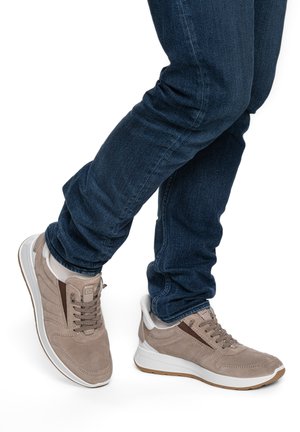 Person trägt beige Sneaker mit weißen Sohlen und blauer Jeans, steht mit einem Bein über dem anderen auf weißem Hintergrund.