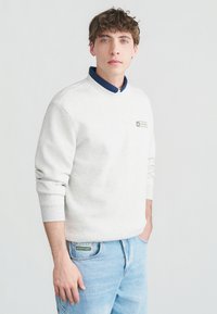 Νεαρός άνδρας με κατσαρά μαλλιά, φοράει ανοιχτό γκρι φούτερ πάνω από μπλε πουκαμίσα και ανοιχτό μπλε τζιν, στέκεται με το ένα χέρι στην τσέπη.