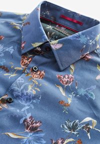 Chemise bleue à boutons avec motif floral multicolore, col ouvert et boutons noirs, présentée pliée et en gros plan.