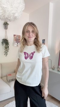 Cămașă albă din bumbac cu un design grafic al unei fluturi violet în centru. Asortată cu pantaloni negri, pe un fundal interior deschis.