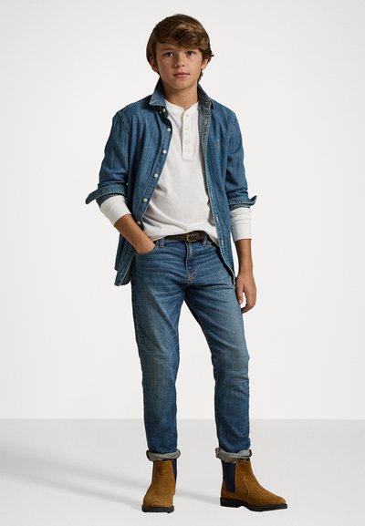Denimjacka över en vit henley, kombinerat med blå jeans och bruna ankelstövlar. Outfiten har uppvikta ärmar och en avslappnad passform.