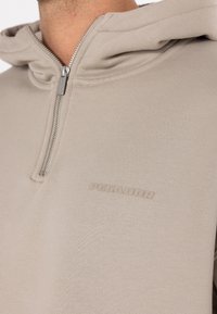 Beige zip-op hoodie med en glat tekstur, kængurulomme og præget logo på brystet. Enkel, funktionel design med hætte.