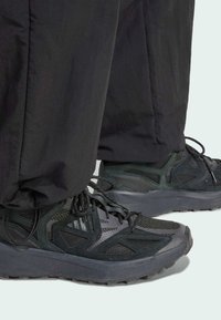 Sapatilhas desportivas pretas com malha texturizada e sobreposições sintéticas. Apresentam uma sola grossa e atacadores, combinadas com calças pretas largas.