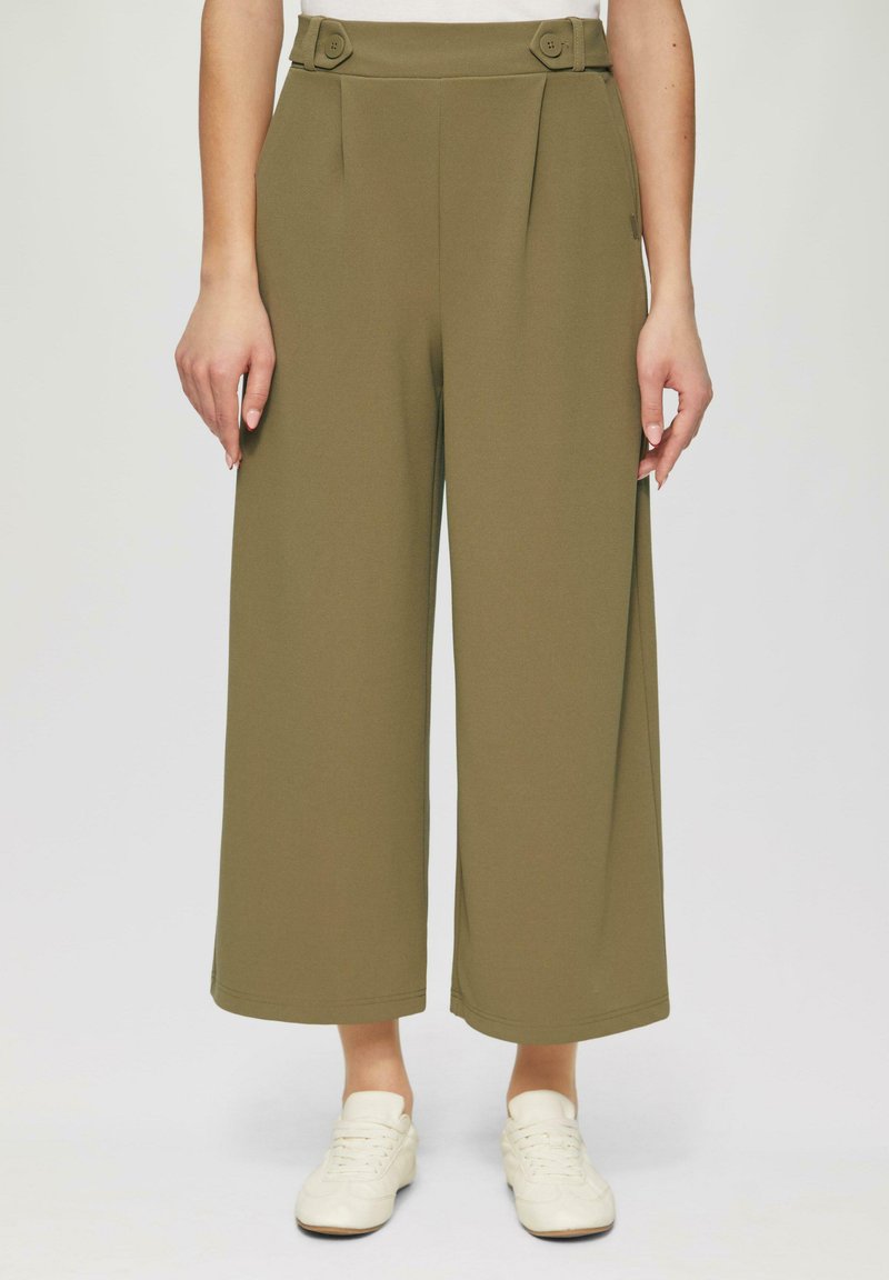 Olijfgroene wijde broek met plooien bij de taille, voorzien van knoopaccenten en een gladde stoftextuur, gecombineerd met witte sneakers.